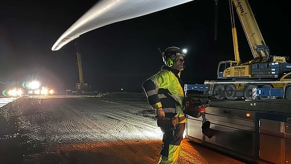 Ein Mann mit Bauarbeiterkleidung und einem Steuergerät steht bei Nacht unter einem Windkraft-Rotorblatt