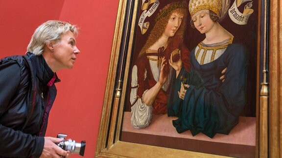 Eine Frau betrachtet ein mittelalterliches Gemälde an der Wand, das zwei eng beieinander sitzende Personen zeigt.