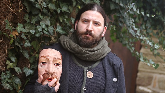 Ein Mann vor einer Mauer mit Efeu hält eine Maske.