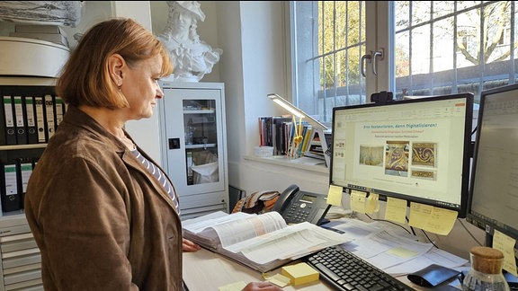 Eine Frau steht vor einem Computer, hält die Maus in der Hand und schaut auf dem Bildschirm.