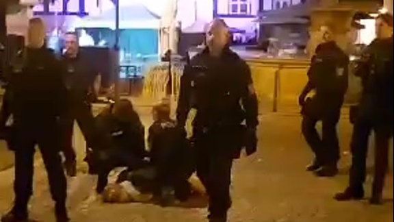 Polizeibeamte stehen auf einem Marktplatz und halten eine Frau am Boden fest.