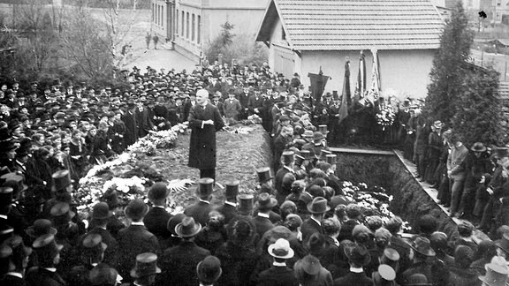 Auf einem historischen Foto sind viele Menschen bei einer Beerdigung zu sehen und ein Redner 