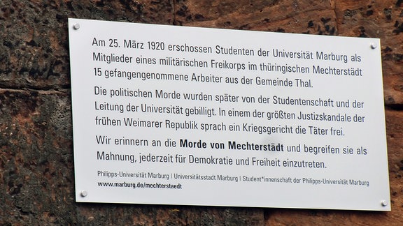An einer Mauer ist eine moderne Tafel zu sehen, auf der die Uni Marburg an die Mordtat von Studenten der Uni in Thüringen erinnert