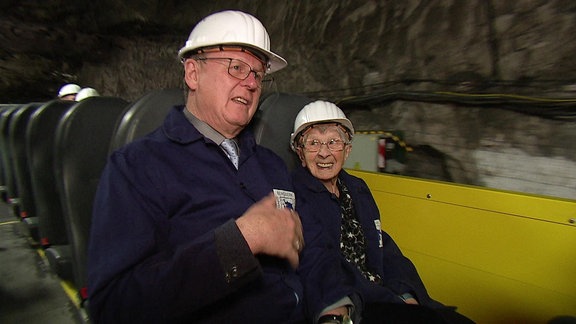 Eine alte Frau und ein Mann sitzen in einem Wagen in einem Bergwerk. Beide tragen Helm.