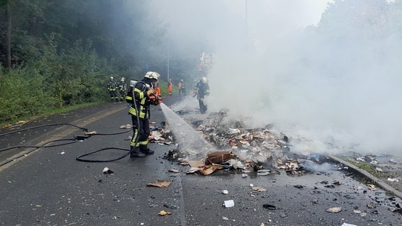 Ein Feuerwehrmann löscht einen rauchenden Berg Papiermüll. Im Hintergrund sind andere Einsatzkräfte und ein Müllauto. 