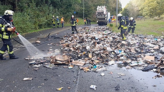 Ein Feuerwehrmann löscht einen rauchenden Berg Papiermüll. Im Hintergrund sind andere Einsatzkräfte und ein Müllauto. 