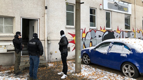 Polizisten stehen vor einem Gebäude, daneben ein Auto.