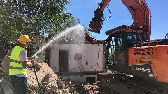 Bauarbeiter in Warnweste neben Bagger spritzt Wasser mit Schlauch auf Haus, das abgerissen wird