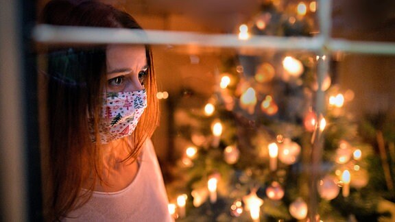 Eine Frau mit Mund-Nasenschutz-Maske schaut sehnsüchtig aus dem Wohnzimmerfenster, innen brennen die Kerzen des Weihnachstbaumes.