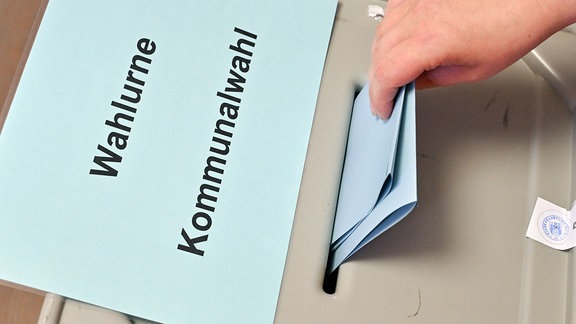 Eine Person steckt ihren Stimmzettel in eine Wahlurne mit der Aufschrift «Wahlurne Kommunalwahl». 