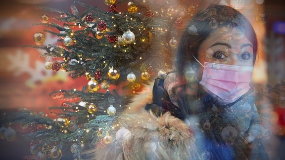 Eine junge Frau mit Nasen-Mundschutz vor einem geschmückten Weihnachtsbaum.