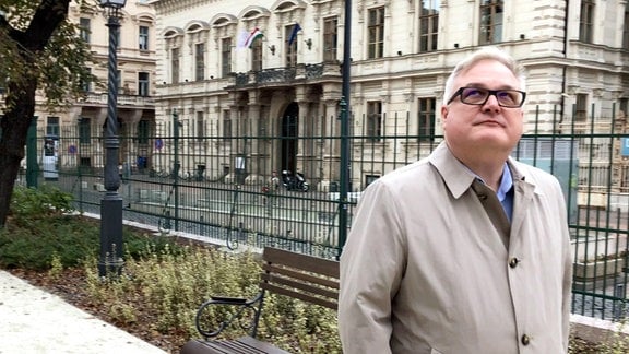 Mann mit grauen Haaren, Mantel und Brille läuft vor einem Palais entlang.