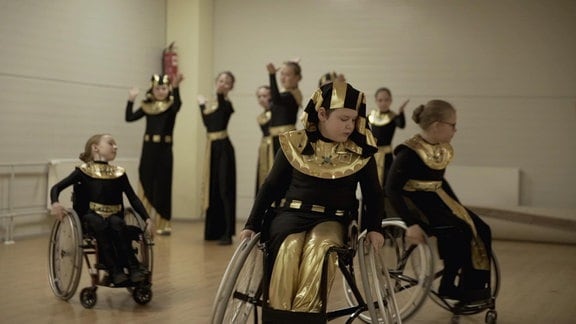 drei Kinder in Rollstühlen und schwarz-goldenen Gewändern bei einer Tanzstunde.
