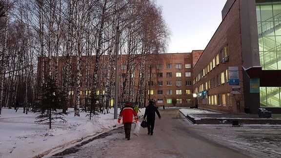 Drei Personen in Anoraks laufen auf einem teilweise vereisten Fußweg. Links ist eine schneebedeckte Fläche zu sehen, auf der viele Birken stehen. Im Hintergrund ist ein roter unscheinbarer Neubau. Bildunterschrift: Das Semaschko-Krankenhaus in Nischni Nowgorod.