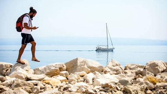 Tourist mit Rucksack geht auf Steinmole entlang, im Hintergrund ein kleines Segelboot.
