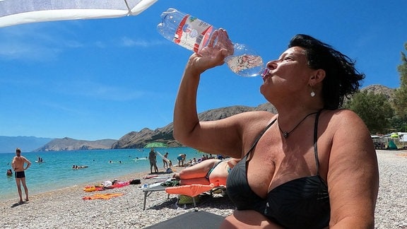 Frau am Stran trinkt aus Wasserflasche.