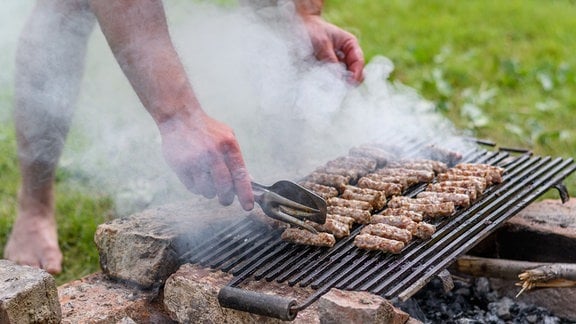 Eine Hand dreht mit einer Grillzange Čevapčići auf einem Grill.