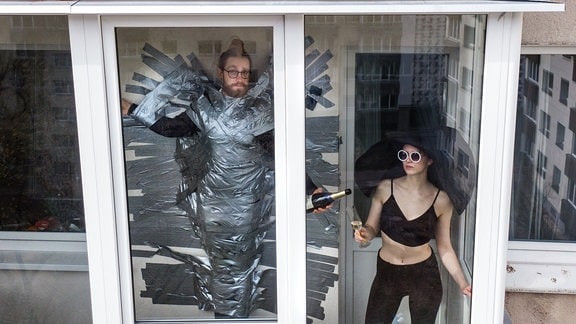 Ein Mann mit Glebestreifen fixiert am Fenster und eine Frau im Kostüm