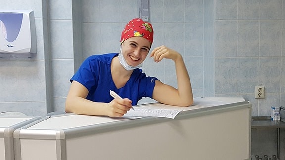 Eine junge Frau in blauem Krankenschwester-Kittel mit roter Haube steht hinter einem Tresen und hält einen Stift in der Hand. Sie lächelt in die Kamera. Hinter ihr stehen Regale mit Krankenhausutensilien. Bildunterschrift: Maria Görlitz während Ihres Freiwilligendienstes in Nischni Nowgorod.