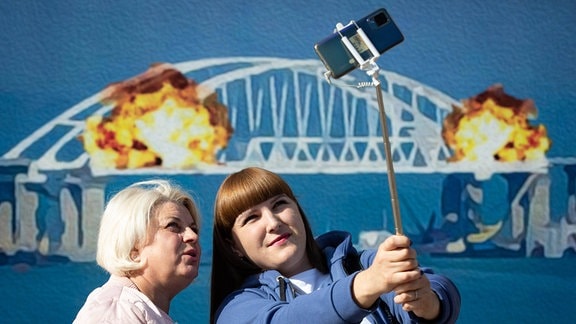 Kiew, Ukraine: Frauen machen ein Selfie mit einem Briefmarkenmodell, auf dem die brennende Krimbrücke im Hintergrund abgebildet ist.