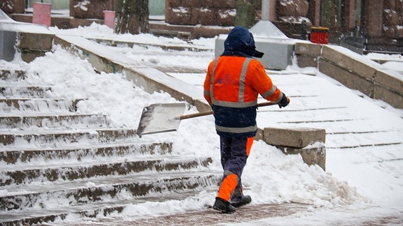 Ein Mann in orangefarbener Weste und Schneeschaufel vor einer Treppe