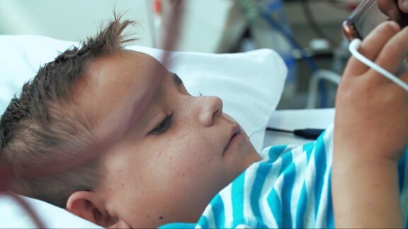 rumänsicher Junge liegt in Klinikbett zur Dialyse und schaut aufs Handy