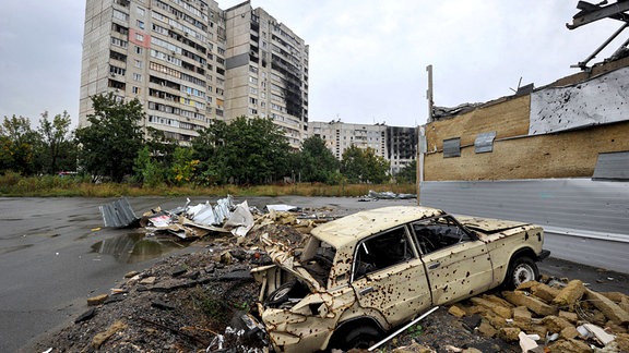 Ein zerstörtes Auto vor dem Hintergrund stark beschädigter Wohngebäude, die durch den Beschuss der russischen Armee beschädigt wurden
