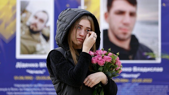 Eine Frau mit Tränen in den Augen und Rosen in der Hand, im Hintergrund Bilder von Soldaten