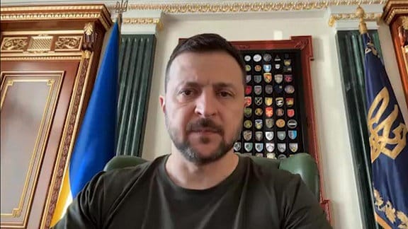 Wolodymyr Selenskyj sitzt auf grünem Sessel und schaut nach unten in die Kamera, im Hintergrund Flaggen