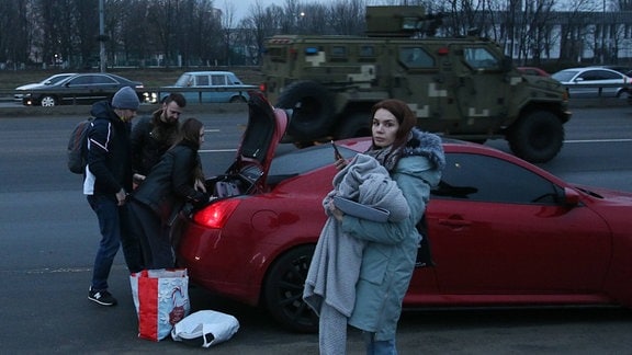 Menschen laden ihre Sachen in ein Auto, während ein Militärfahrzeug auf einer Straße in Kiew, Ukraine, vorbeifährt.