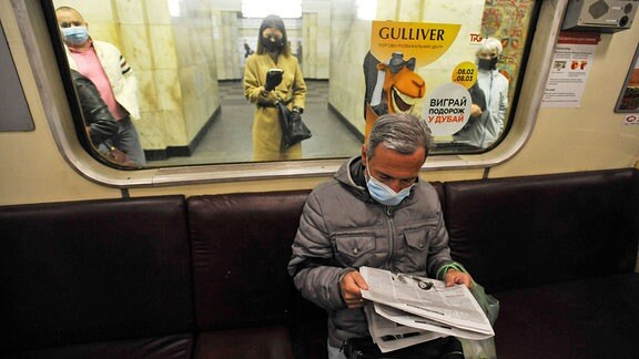 Ein Mann, der in der U-Bahn sitzt und Zeitung liest, am Bahnsteig warten Menschen