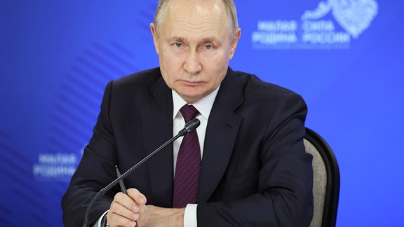 Wladimir Putin sitzt zuhörend vor einem Mikrofon