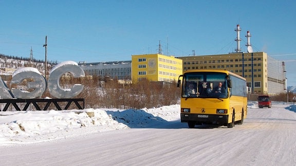 Ein gelber Bus fährt auf einer weiten, schneebedeckten Straße am Gebäude des Bilibino Atomkraftwerks vorbei.