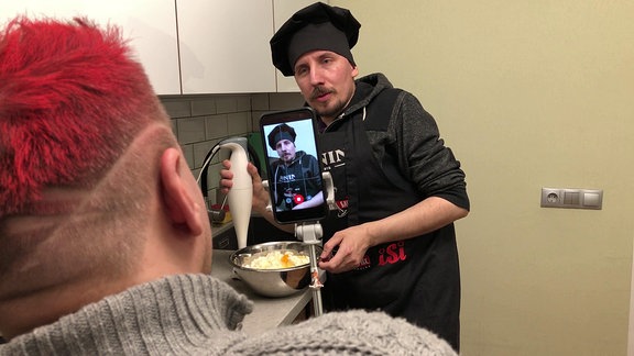 Ein Smartphone auf einem Selfie-Stick filmt einen Mann mit schwarzer Kochmütze und Schürze, der auf dem Display und im Hintergrund zu erkennen ist. Im Vordergrund  sind Kopf und Schultern des Mannes zu sehen, der den Koch filmt.