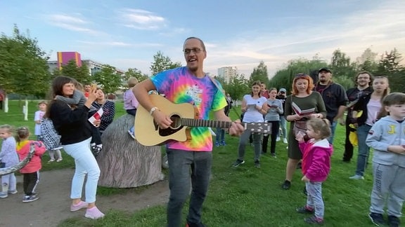 Mann in buntem Shirt spielt Gitarre in einem Park, umringt von singendne Erwachsenen und Kindern.
