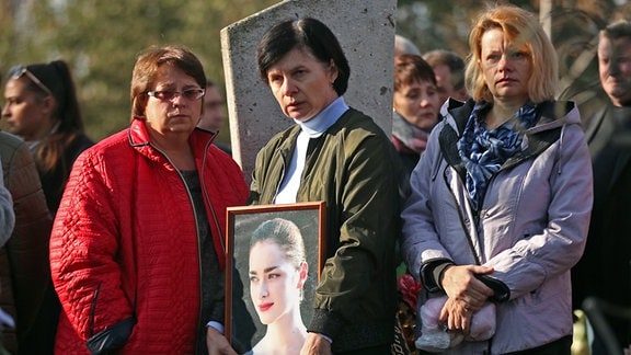 Drei frauen jalten das gerahmte Bild einer jungen Frau bei einer Beerdigung.