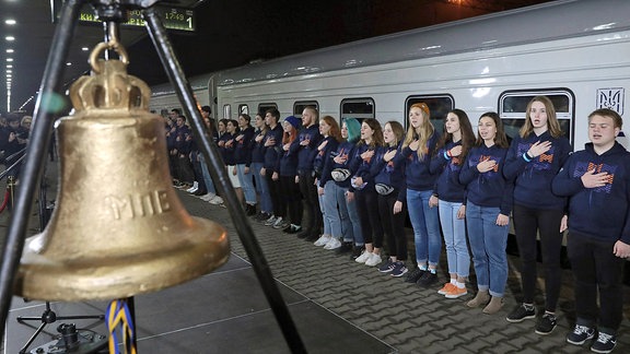 Junge Menschen stehen auf einem Bahnsteig neben einem Zug, daneben eine Glocke