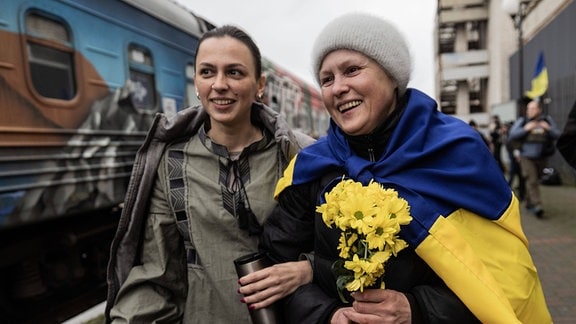 Zwei Frauen stehen an einem Bahnsteig und lächeln.