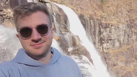 Ein Student mit Sonnenbrille lächelt verschmitzt in die Kamera. Im Hintergrund ein Wasserfall.