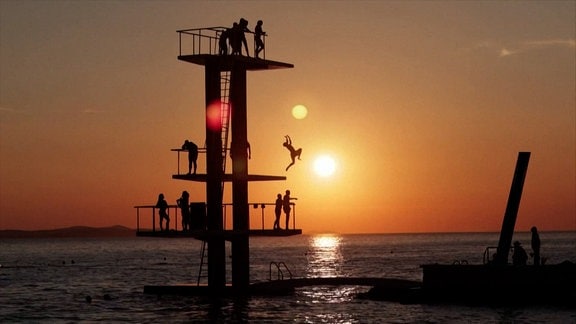 In der Abendsonne ein hoher Sprungturm im Meer. Menschen springen ins Wasser. 