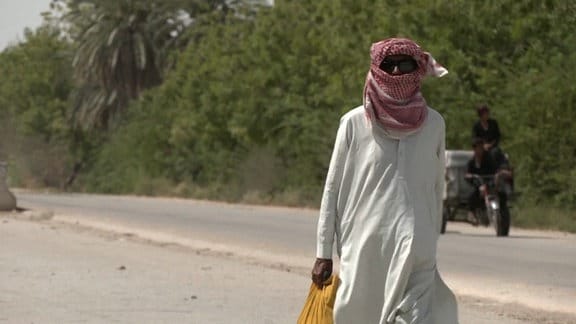 Ein Mann mit einem Kopftuch, der eine staubige Straße entlangläuft. Hinter ihm kommt ei Moped angefahren.  
