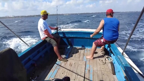 Zwei Fischer nehmen am Hemingway-Angel-Turnier teil. Sie sitzen am Heck eines Fischkutters, und angeln; vor sich blaues Meer mit Sonnenschein.