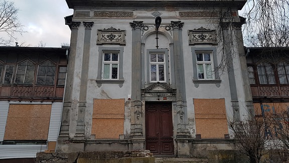 Heruntergekommenes Eingangshaus eines größeren Gebäudes mit neoklassizistischen Säulen, Jugenstilornamenten und zugenagelten Erdgeschoss-Fenstern