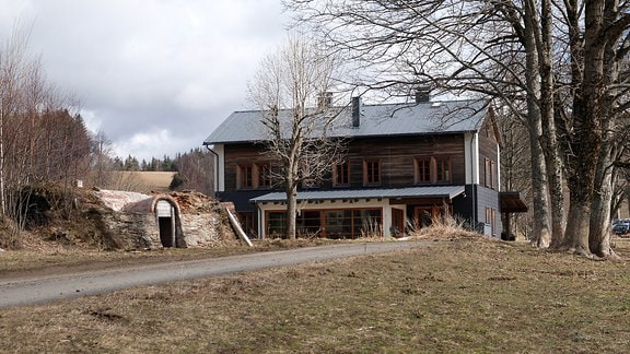 Ein großes, zweistöckiges Bauernhaus mit viel Holzverkleidung steht an einem Waldrand