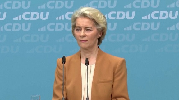 Hintergrund: eine Blaue wand mit dem CDU-Logo (Buschatebn, C, D, U in weiß). Vordergrund: Ursula von der Leyen in einem braunorangenem Sakko mit beiggelben Oberteil. Vor ihr zwei dünne Mikrofone.  Ihr Blick Neutral bis ernst. 