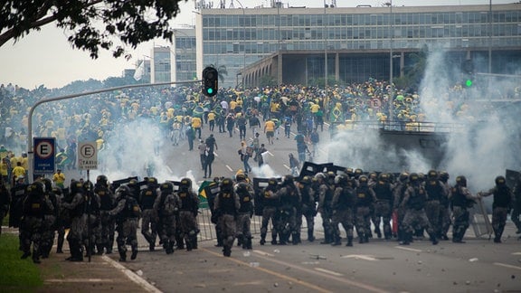 Anhänger des ehemaligen brasilianischen Präsidenten Bolsonaro (hinten) geraten in der Hauptstadt mit Ordnungskräften aneinander, die hinter Absperrungten eine Kette bilden und Tränengasgranaten auf die Demonstranten abfeuern.