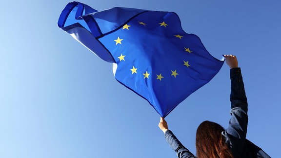 Frau hält Flagge der Europäischen Union vor blauem Himmel im Freien, Blick aus niedrigem Winkel.