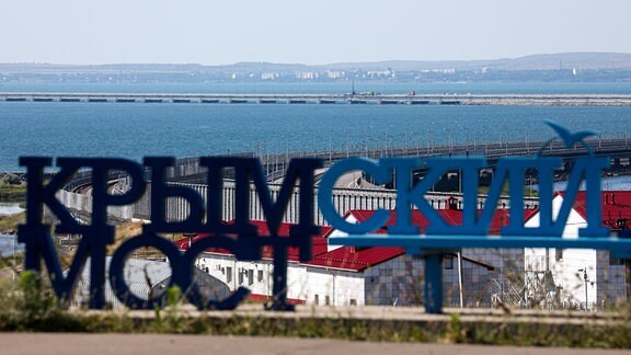 Der Schriftzug «Krim-Brücke» steht auf dem Festland mit der beschädigten Krim-Brücke, die das russische Festland und die Halbinsel Krim über die Straße von Kertsch verbindet, im Hintergrund. 