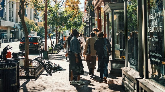 Auf einem Gehweg des Stadtteils Chinatown ist eine Gruppe junger Menschen zu erkennen, die von der Kamera weglaufen und teilweise ein modernes Restaurant mit Glasfassade betreten.