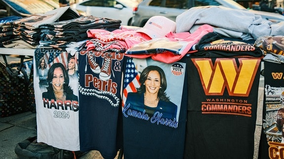 Auf einem Klapptisch am Rand des Gehwegs stapeln sich verschiedene T-Shirts, auf der Hälfte davon ist die demokratische Präsidentschaftskandidatin Kamala Harris zu sehen.